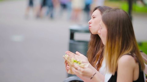 Caucasian women eating hamburger fast food sandwich on the street outdoors. Active girls hungry and eating street food after long walk