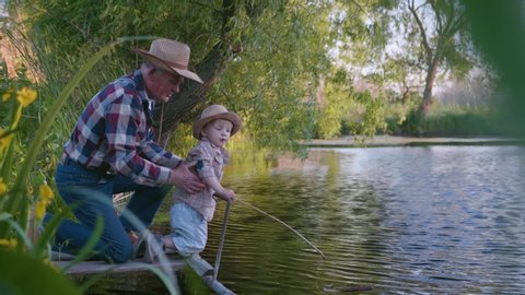 fishermen, caring loving grandfather teaches his little cute grandson to fish with fishing rods in river standing on pier background of reeds and trees