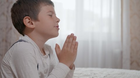 religion, prayer male child trust in God with hope in his heart and with folded together arms standing on wheels near bedstead in apartment, close-up