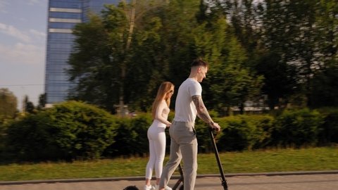 Young beautiful guy and girl in white clothes ride electric scooters in the Park at sunset. Slow motion.