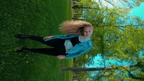 A beautiful girl is grimacing in front of the camera, has lost her curly hair and twists her head. The wide angle of the lens leans along with the blonde. Fun on 4K video in a green park in summer.