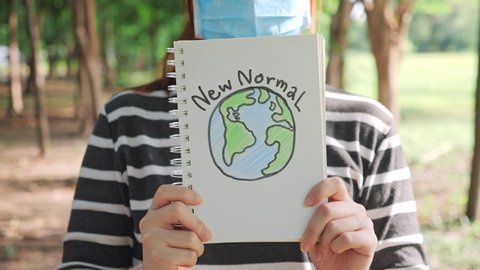 Woman wearing protective mask is holding word "New Normal" is written in a book with the green nature background, New Normal concept. covid-19, coronavirus and new normal lifestyle concept