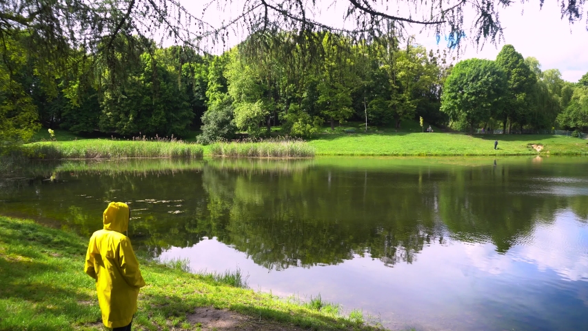 Woman in yellow raincoat walking down to lake in summer park. Girl admires river landscape view relaxing outdoors
