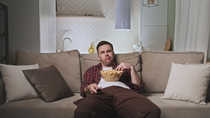 Bored and unhappy man watching TV. 30s male watching TV at night after hard day sitting on couch with big bowl of popcorn. 4K, UHD Royalty-Free Stock Footage #1053836366