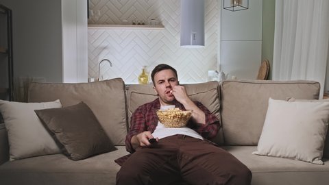 Bored and unhappy man watching TV. 30s male watching TV at night after hard day sitting on couch with big bowl of popcorn. 4K, UHD