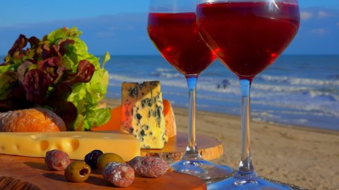 Romantic picnic by the sea with red wine in glasses, various sorts of cheese, bread, lettuce and olives