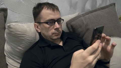 Serious Man in Black Polo T-shirt and Glasses Lying on Couch with Mobile Phone in His Hands, Looking at Cellphone, Touch the Screen, Scrolling and Reading News
