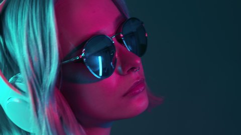 Gorgeous woman with dyed hair listening music in headphones and singing with neon light background. Charming hipster girl dancing with sunglasses