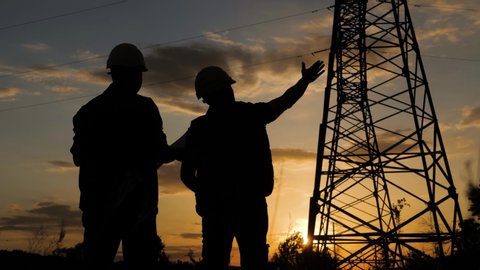 Silhouette of engineers looks at the construction of high-voltage power. Teams engineer looking discussing plan. Two engineer standing on field with electricity towers at sunset.