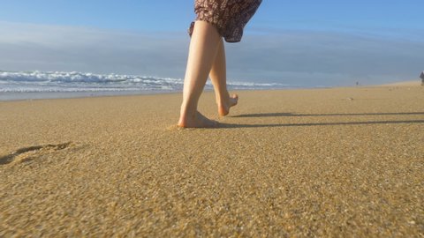 lonely woman legs go along the ocean barefoot on the sand leaving footprints close-up, the wave washes her feet with splashes slow motion