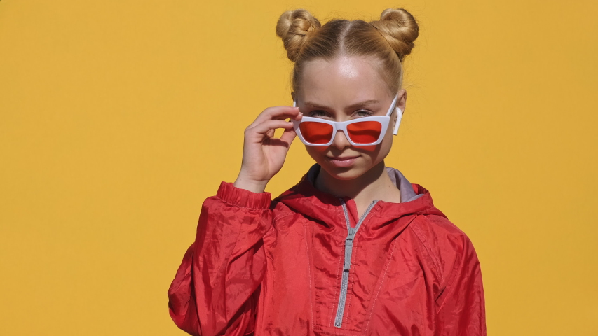 Portrait of happy teen girl blonde in airpods in stylish sunglasses, red hoodie smiles, dances, rhythmically shaking his head camera on yellow background slow motion. Emotions of people. Girl dancing Royalty-Free Stock Footage #1053852029