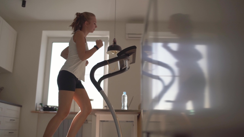 Woman running on running machine at home.Full length profile shot of a fit woman jogging on a treadmill in the kitchen. | Shutterstock HD Video #1053855398