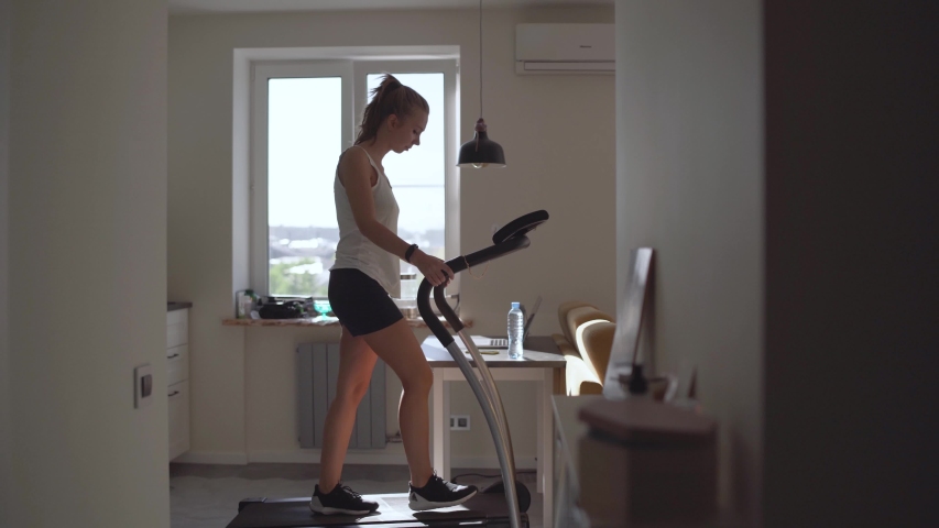 Woman running on running machine at home.Full length profile shot of a fit woman jogging on a treadmill in the kitchen. | Shutterstock HD Video #1053855416