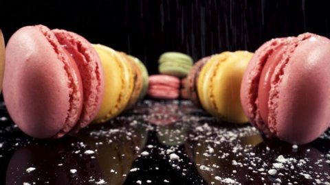 Reverse moving macro view past macarons with powdered sugar falling on black reflective glass, luxury french pastries, sweet sugary food