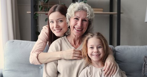 Head shot portrait happy young woman cuddling smiling hoary mature senior mother and adorable little preschool daughter, relaxing together on comfortable sofa, 3 generations warm relations concept.