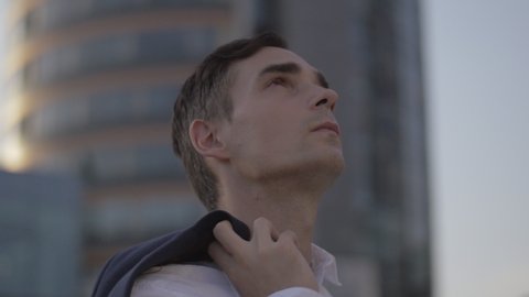 Dynamic shot of young businessman surrounded by office buildings.Handsome man holding a jacket over the shoulder and looking around at the distance in the city.