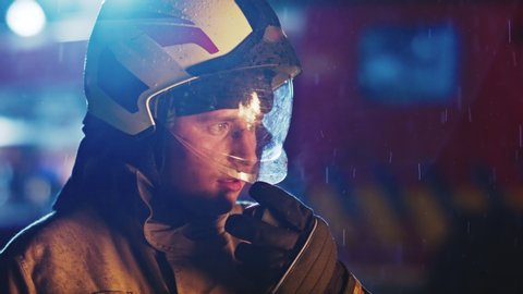 Portrait shot of a fireman speaking on the walkie talkie and the site during the rain. Fire reflection on the helmet. Slow motion.