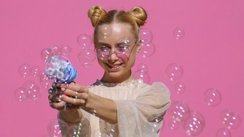 Portrait of happy blonde teen girl in stylish sunglasses with soap bubbles smiling releases toys from gun with pleasure on pink background in summer slow motion. Emotions, joy. Childhood