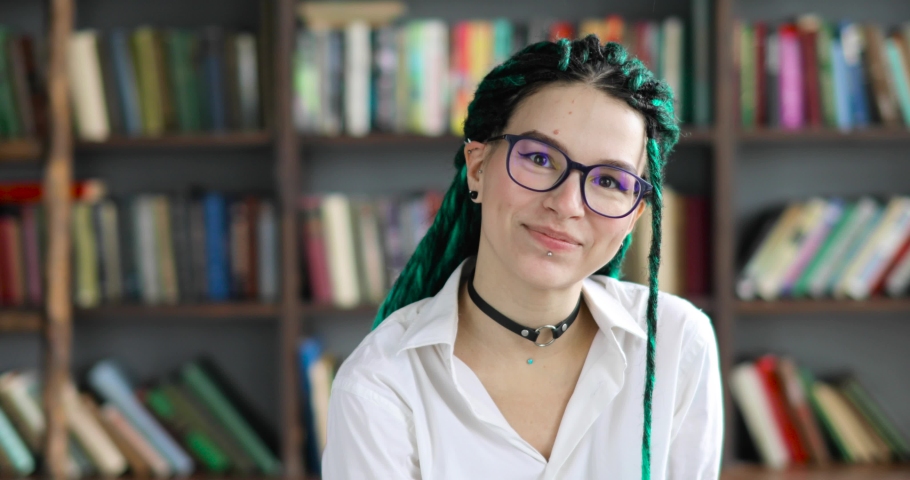 Pretty girl student with green dreads hairs in glasses in library looking at camera. Portrait of smiling young girl with green dreadlocks in white blouse on bookshelves background. Studying concept. Royalty-Free Stock Footage #1053877967