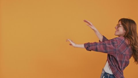 Red-haired adolescent girl in checkered shirt and glasses. She is dancing, showing tongue and smiling while moving along orange background. Close up