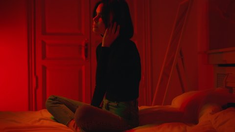 Young woman dancing to beat with her eyes closed in red neon light. Cute girl enjoying music in wireless headphones late night at home in bedroom. Teen girl sitting on bed in her room