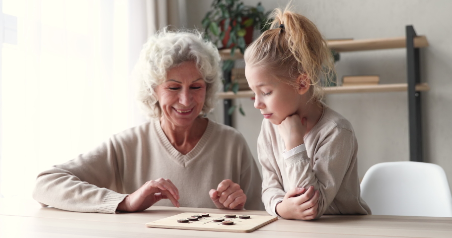Smiling middle aged senior hoary grandmother playing wooden draughts on checkerboard with cute focused little preschool granddaughter, enjoying hobby boardgame pastime together in living room. Royalty-Free Stock Footage #1053881159