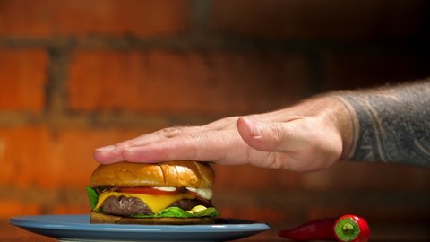 Male hand in tattoos kneads a burger, cheese and sauce flow out. Juicy tasty burger lies on a plate with red chili peppers. Unhealthy foods in self-isolation due to coronavirus
