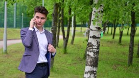 Young attractive male businessman in a gray suit walks in a green park and speaks on the phone. Smiling in a positive mood.