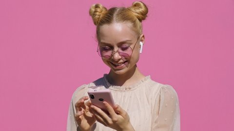 Portrait of happy teen blonde girl in airpods in pink glasses listens to music on smartphone, smiles, dances, shaking her head rhythmically against pink background of slow motion in summer. Emotions Video Stok