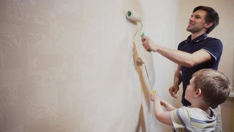 Dad and son with smile paint beige wallpaper on the wall with white paint with a large and small roller Stock Video