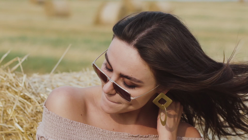 Portrait of happy woman in sunglasses with blowing hair smiling in field at haystack. Slowly