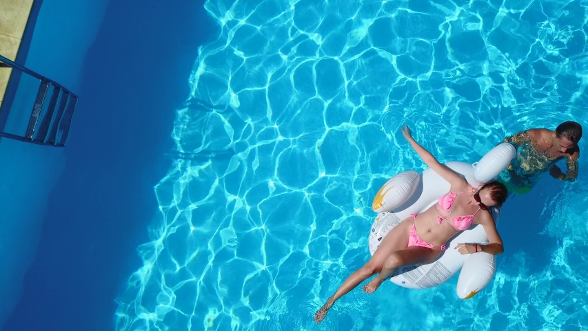 Aerial. Friends chilling in swimming pool with inflatable flamingo, swan, mattress. Happy young people bathe on floating mattresses in luxury resort. View from above. Girls in bikini sunbathing in sun | Shutterstock HD Video #1053899843