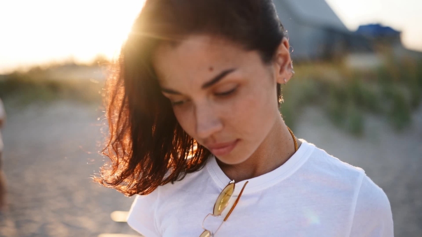 Portrait of young woman looking at camera standing on the beach with a sea on background. Attractive girl in white t-shirt raises eyes. Wind blows airy brown hair shining in sun flares.