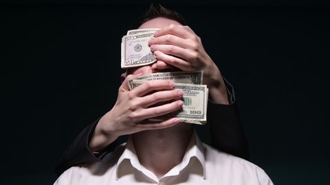 Unknown hands from the dark tightly cover mouth and eyes with money to a young man in a white shirt. The man is silenced and closed his eyes to what is happening