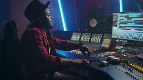 Energetic Male Audio Engineer / Producer Working in Music Recording Studio, Mixing Tracks on Control Desk and Software to Create Hit Song Track. Black Artist Musician Enter Studio and Sits at Workdesk