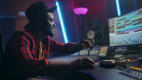 Energetic Audio Engineer / Producer Working in Music Recording Studio, Mixing Tracks on Control Desk and Software to Create Distinctive Modern Sound of Hit Song. Portrait of Black Artist Musician.