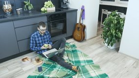 Man in glasses sitting on floor in kitchen with laptop computer, eating apple and rubbing eyes after hard working day. Male freelancer using notebook and thinking about solution of problem at work
