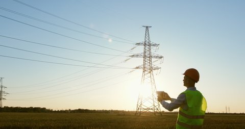 An energy engineer in special clothes inspects a power line using data from electric sensors on a tablet.