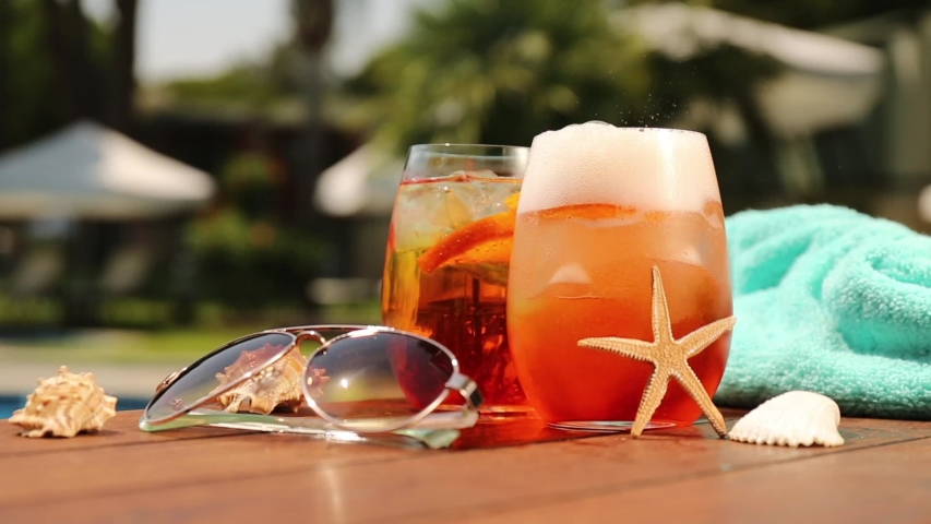 Glasses of aperol spritz or negroni cocktail with seashells, towel and sunglasses at the summer patio. Vacation, summer, holiday, luxury resort concept | Shutterstock HD Video #1053921032