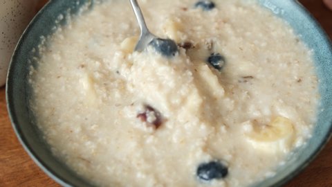 Stirring breakfast oatmeal porridge with bananas and blueberries. Eating healthy vegetarian food, breakfast cereals