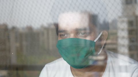 A shot of a young man through the glass window is wearing a green protective face mask and looking out. A distressed or worried patient is quarantined during Coronavirus or COVID 19 epidemic  