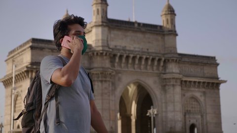 A young attractive man wearing protective face mask and talking or speaking on mobile phone or smartphone in front of historic landmark Gateway of India amid coronavirus or Covid19 epidemic