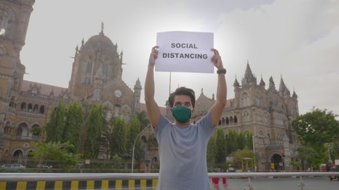 A young man wearing face mask standing and holding a placard with message 'Social Distancing' during city lockdown amid coronavirus corona against historic CSTM or VT terminus station