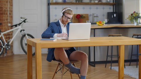 Portrait of funny young businessman wearing no pants and listening to music from headphones sitting at desk and working on laptop during self-isolation