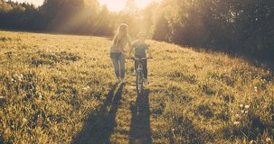 Mother teaching son to ride bicycle. Happy cute boy in helmet learn to riding a bike in park on green meadow in summer day at sunset time. Family weekend. 4K video Slow motion
