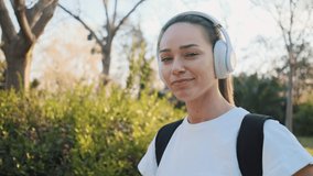Tracking around portrait shot of beautiful girl in headphones with backpack smiling on camera on the way to training in the park. Morning Sun flare 