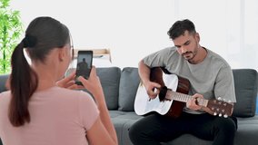 4K Young couple playing the guitar at home while his girlfriend is recording video with smartphone and enjoying music