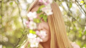 Video clip, blur effect and light flare, a young blonde woman with long hair in a long dress walks in a blossoming apple orchard on a sunny day in spring