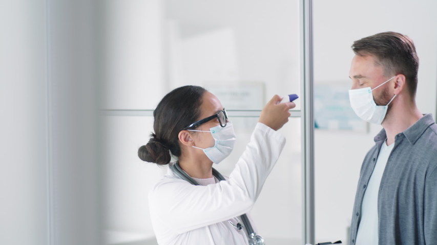 Female doctor in protective face mask scanning forehead of male patient with digital infrared thermometer and writing down measurement on clipboard during coronavirus testing in clinic | Shutterstock HD Video #1053952772