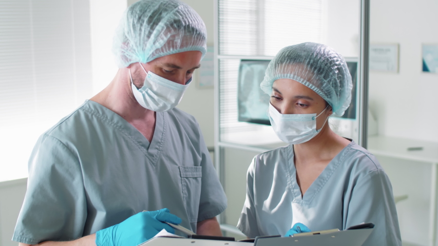 Male and female doctors in protective masks, disposable hats and gloves discussing something on clipboard while working together in hospital during coronavirus pandemic Royalty-Free Stock Footage #1053953186